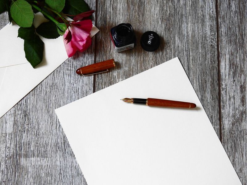 Fotografia di un quaderno bianco con una penna sopra e una rosa accanto